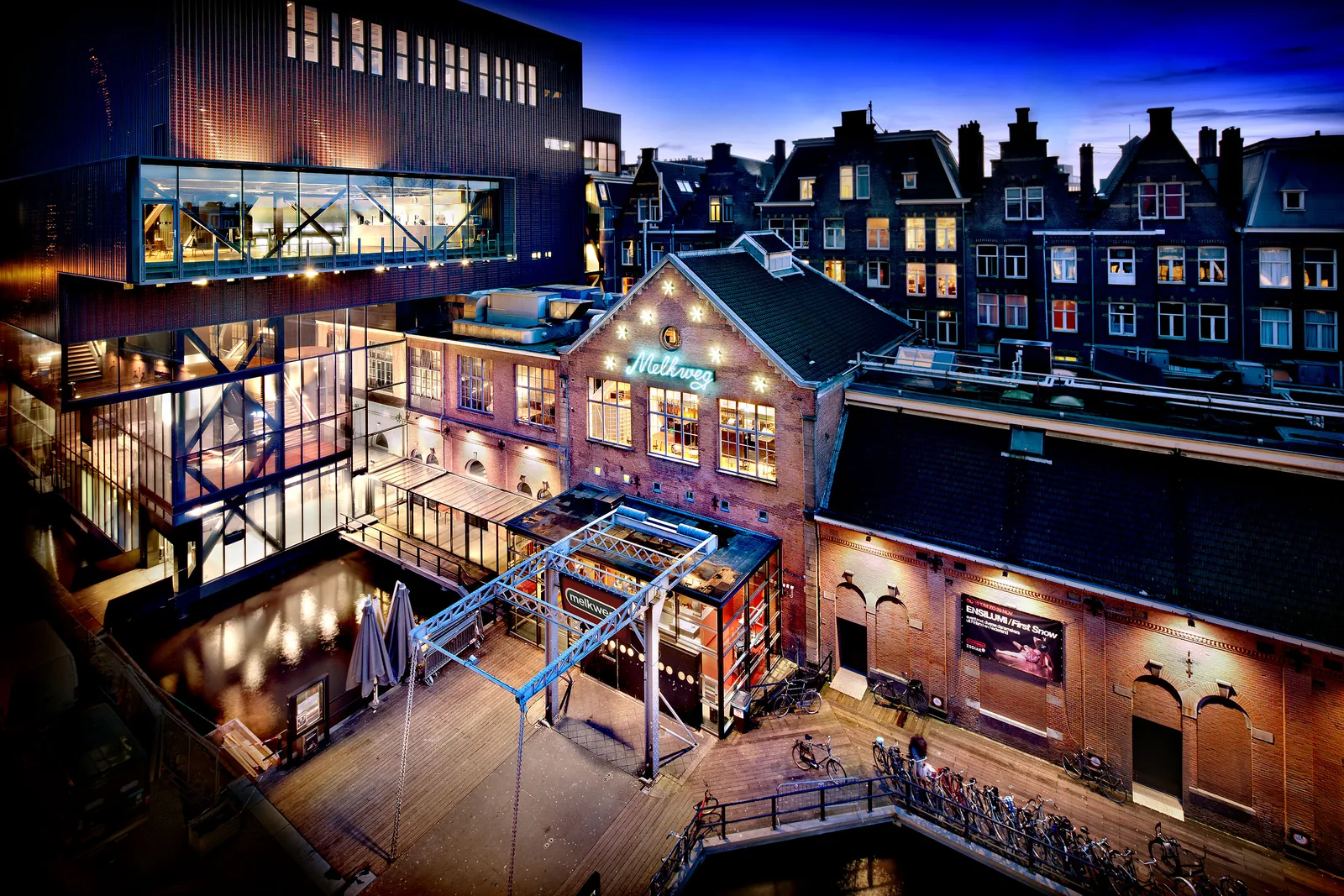 Melkweg at night, green neon on the brick gable, a glass hall to the left, canal houses and bicycles along the water.