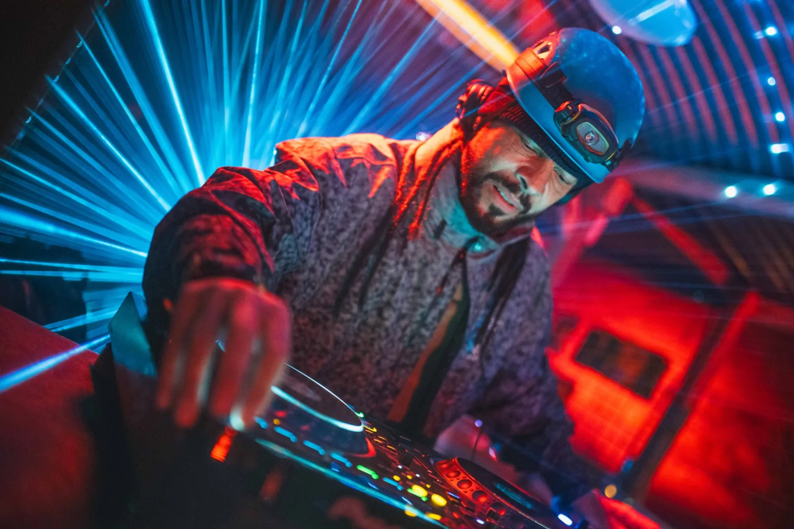 Roni Size at the decks in a blue climbing helmet with the headlamp still clipped on, headphones over a black beanie, right hand on the jog wheel, blue lasers and red light.