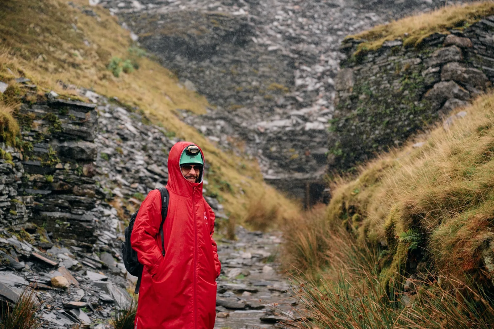 A person in a long red cagoule, green cap and headlamp, standing in rain on a broken-slate path between quarry walls above Cwmorthin.