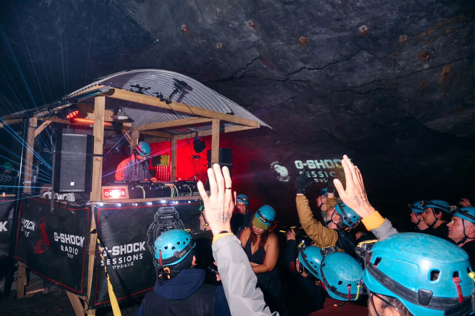 A timber DJ booth with a corrugated roof inside a slate cavern, teal climbing helmets in the crowd, hands raised, G-SHOCK Sessions projected on the rock