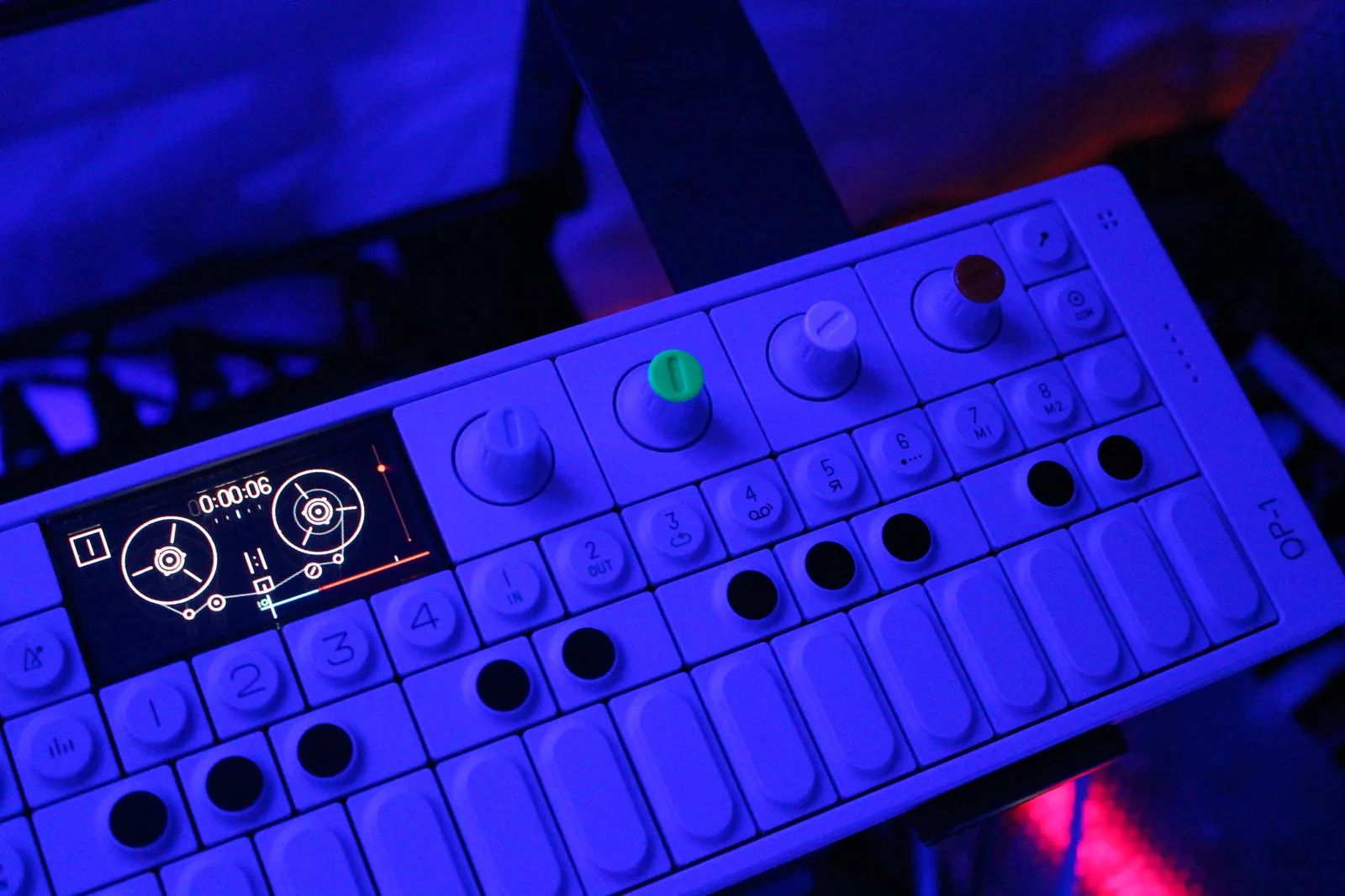 A white Teenage Engineering OP-1 under blue light, tape reels on the screen, colored knobs above the keys.