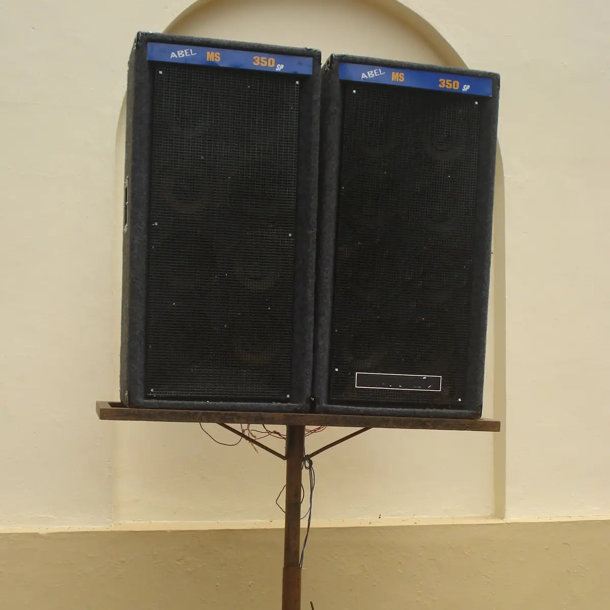 A lone pair of black loudspeakers on a tripod stand against a pale wall, no one around.