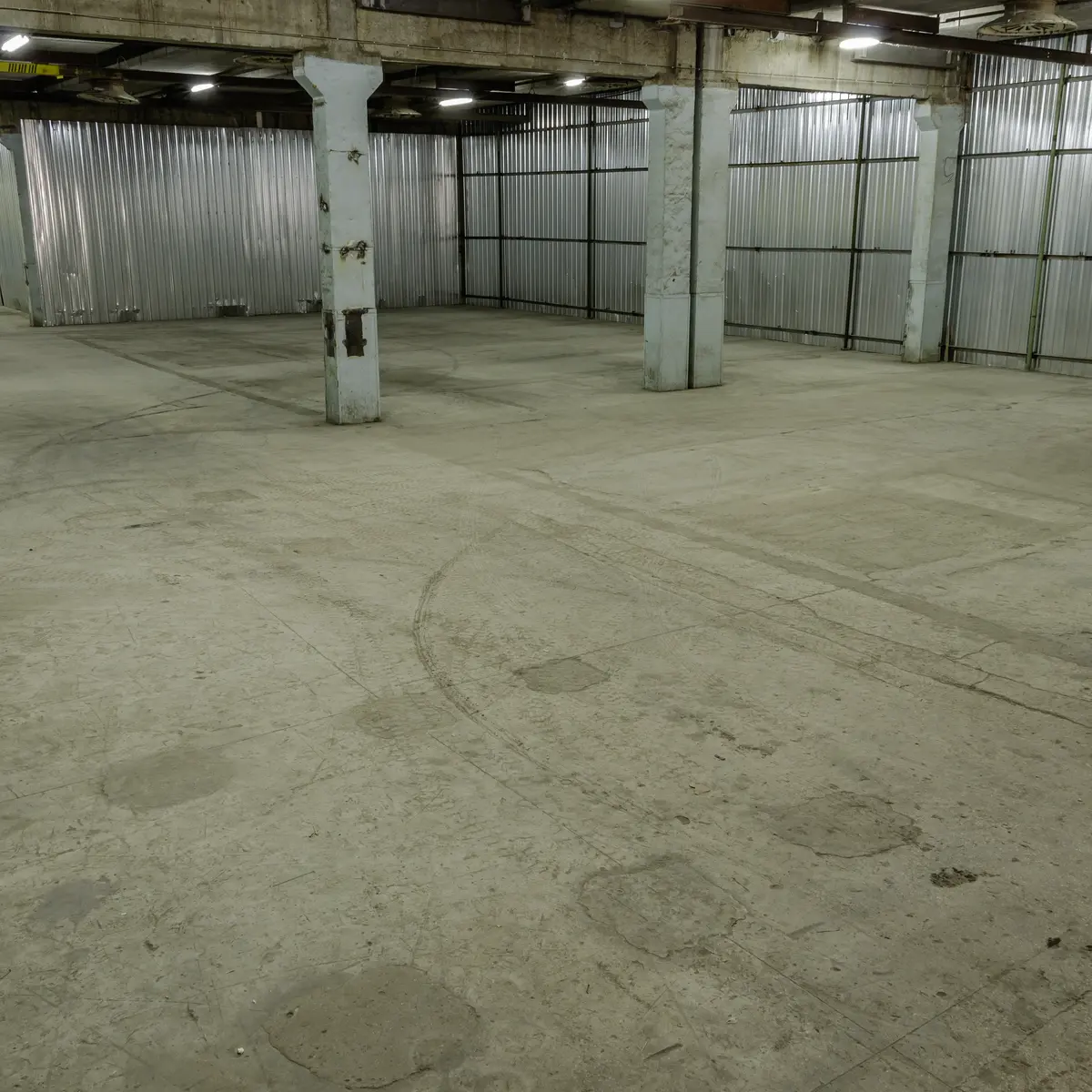 An empty warehouse with concrete pillars, corrugated metal walls and a scuffed bare floor under strip lights.