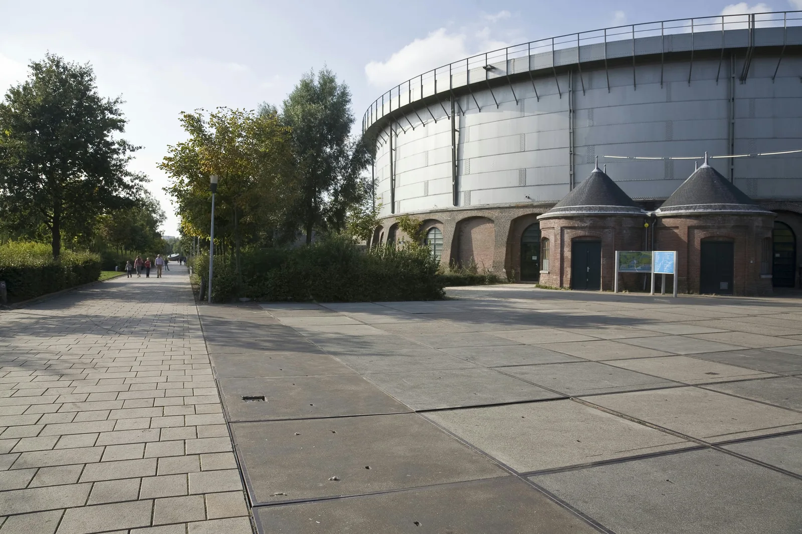 The round silver gas holder at Westergasfabriek in daylight, brick arches along the base, two pointed brick porches, an empty paved yard.