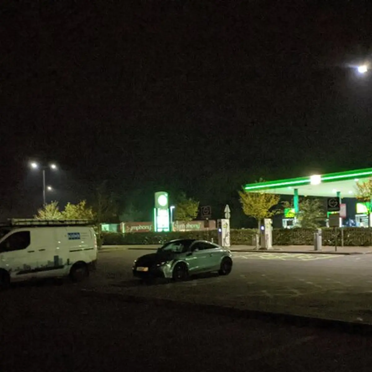 A motorway service station forecourt glowing green under streetlights at night, a few parked cars, nobody in view.